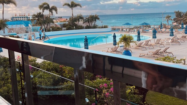 A pool area with lounge chairs and umbrellas, a clear view of the ocean, and a building with a blue and white facade in the background.