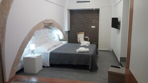 A modern hotel room with a curved ceiling, a bed with a dark bedspread, a bedside table with a lamp, a flat-screen TV mounted on the wall, and a small seating area with a chair and a table.