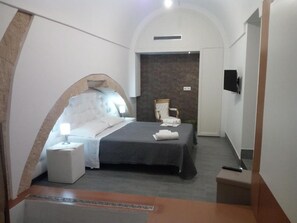 A modern hotel room with a curved ceiling, a bed with a dark bedspread, a bedside table with a lamp, a flat-screen TV mounted on the wall, and a small seating area with a chair and a table.