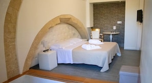 A hotel room with a large arched stone wall, a bed with white linens, a bedside table with a lamp, and a small round table with a chair. - Hotel San Giorgio (Modica)