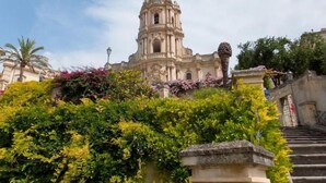 A historic building with a prominent bell tower, surrounded by lush greenery and a stone staircase.