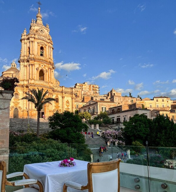 A terrace with a table set for dining, overlooking a historic building with a bell tower and steps leading up to it.