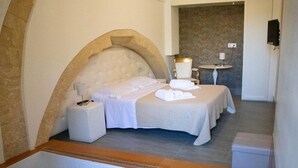 Une chambre d’hôtel avec un grand mur en pierre en arc de cercle, un lit recouvert de draps blancs, une table de chevet avec une lampe et une petite table ronde avec une chaise.