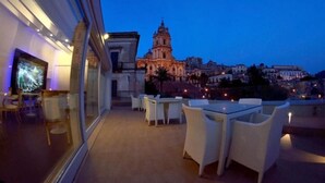 An outdoor terrace with white wicker furniture, a glass wall, and a view of a historic building at dusk.
