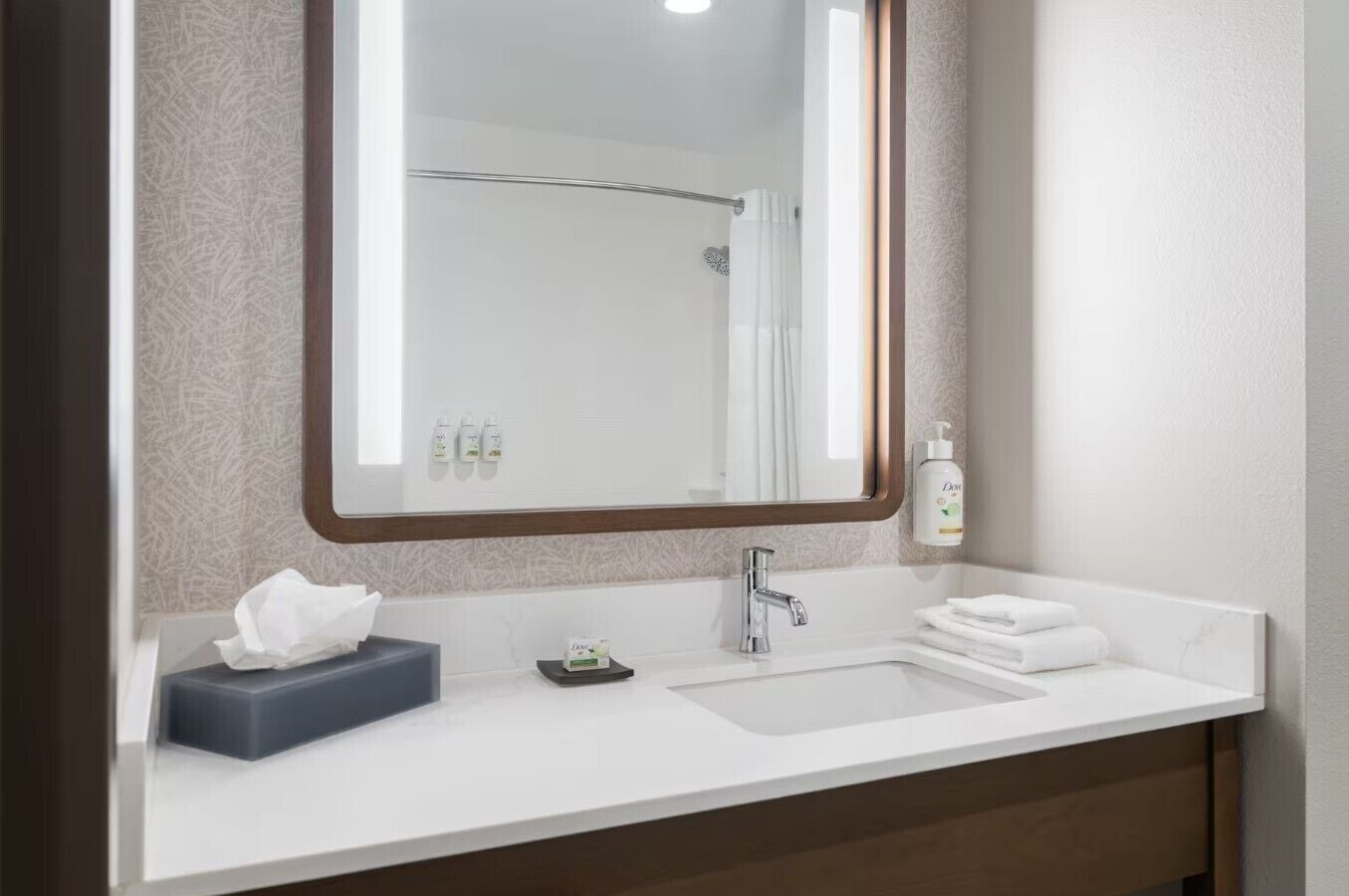 Casa de banho de hotel com uma pia branca, um espelho, um dispensador de toalhas e uma caixa de lenços.