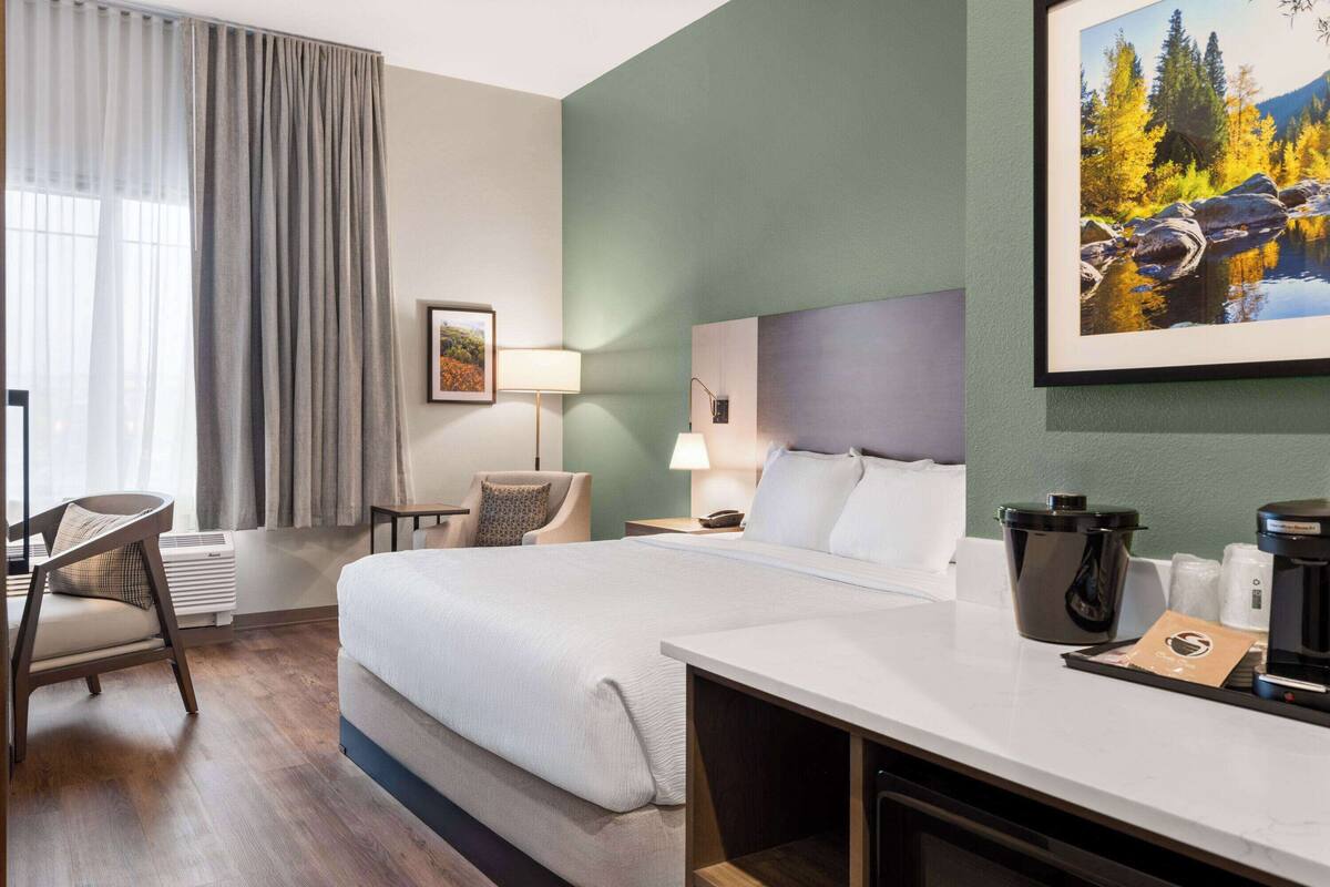 Une chambre d’hôtel avec un grand lit, un bureau avec une machine à café, une chaise, une lampe et un tableau représentant un paysage forestier.