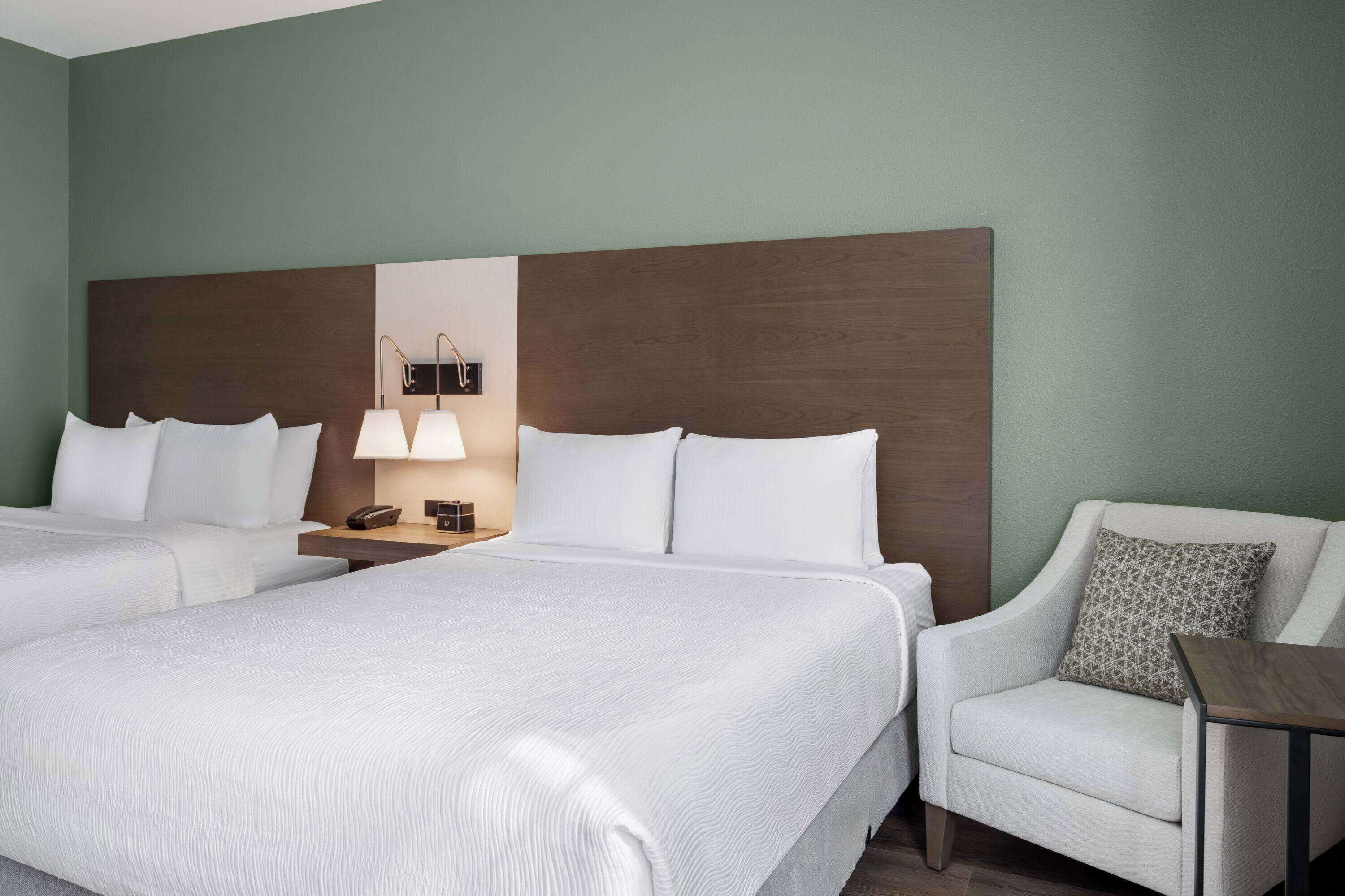 Ein Hotelzimmer mit zwei Betten, einem Nachttisch mit Lampe, einem Sessel und einem kleinen Tisch.