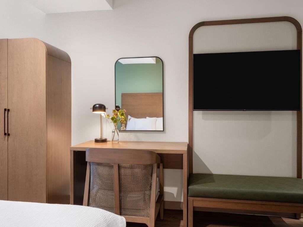 Une chambre d’hôtel avec une armoire en bois, un bureau avec une lampe, un miroir et un banc rembourré de couleur verte.