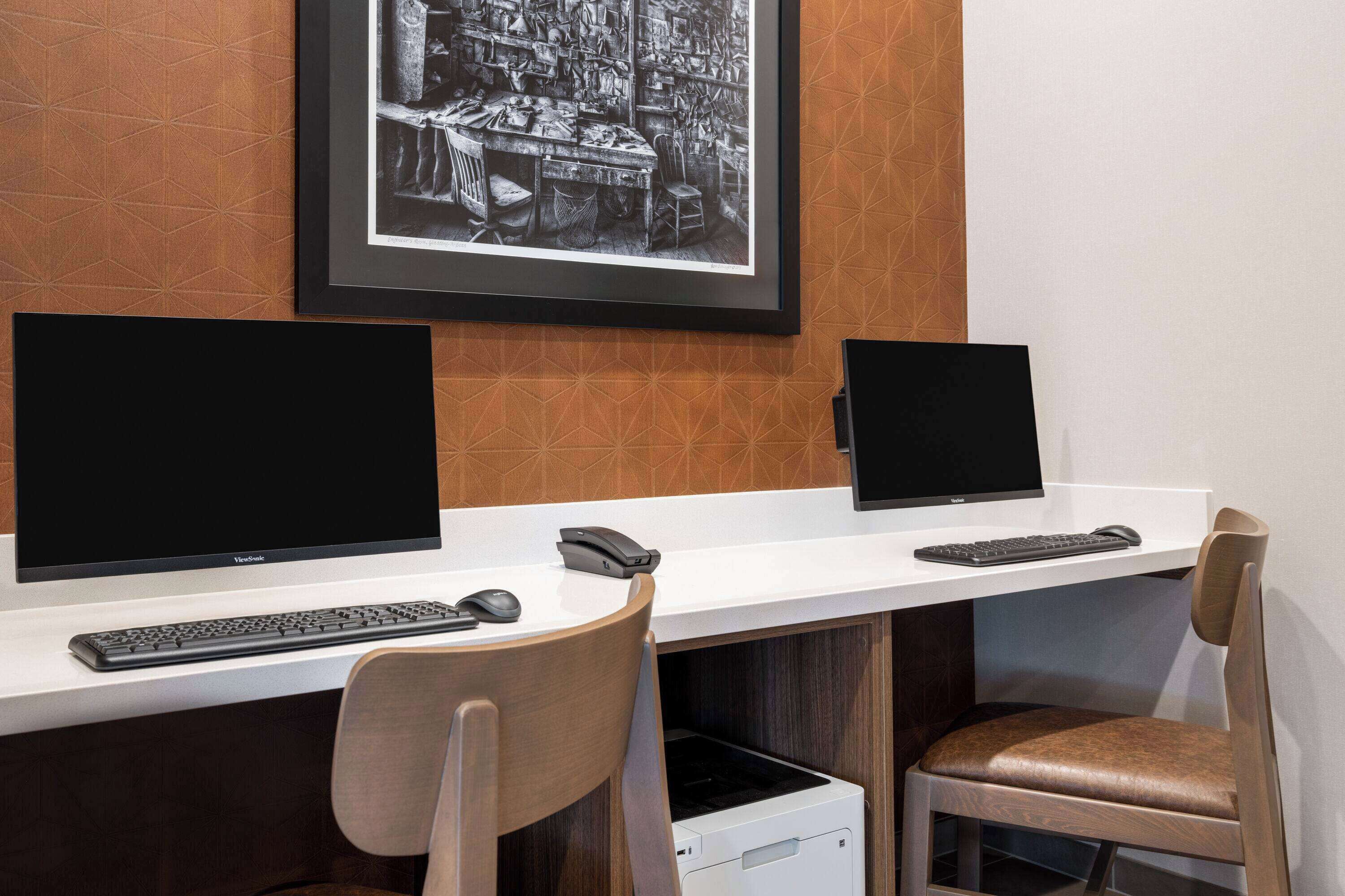 Ein Arbeitsplatz mit zwei Schreibtischen, jeweils ausgestattet mit einem Computermonitor, Tastatur und Maus. Zwei Stühle gehören zur Einrichtung, einer mit einer Holzlehne und der andere mit gepolstertem Sitz. Über den Schreibtischen hängt ein gerahmtes Schwarz-Weiß-Foto.