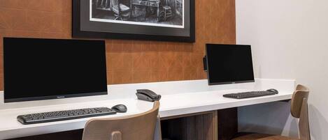A workspace with two desks, each with a computer monitor, keyboard, and mouse. There are two chairs, one with a wooden backrest and the other with a cushioned seat. A framed black and white photograph is displayed on the wall above the desks.