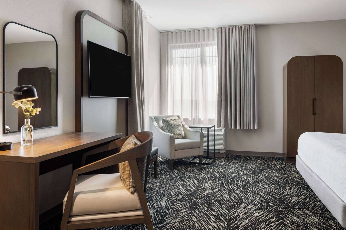 Une chambre d’hôtel avec un bureau en bois, une chaise, un lit, une télévision, une petite table avec une chaise et une fenêtre avec des rideaux.