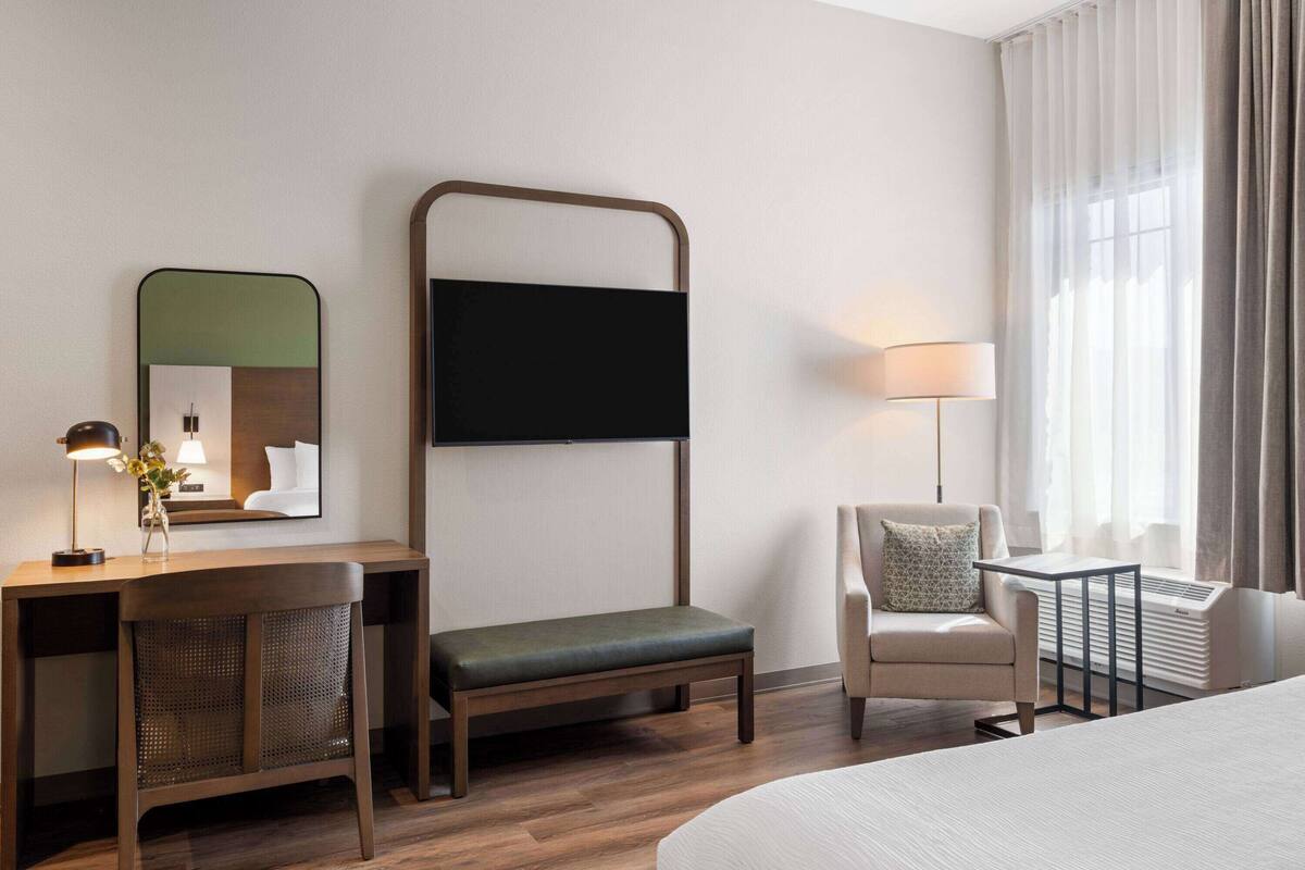 Une chambre d’hôtel avec un lit, un bureau doté d’une lampe, une chaise, un banc, un miroir et une fenêtre avec des rideaux.