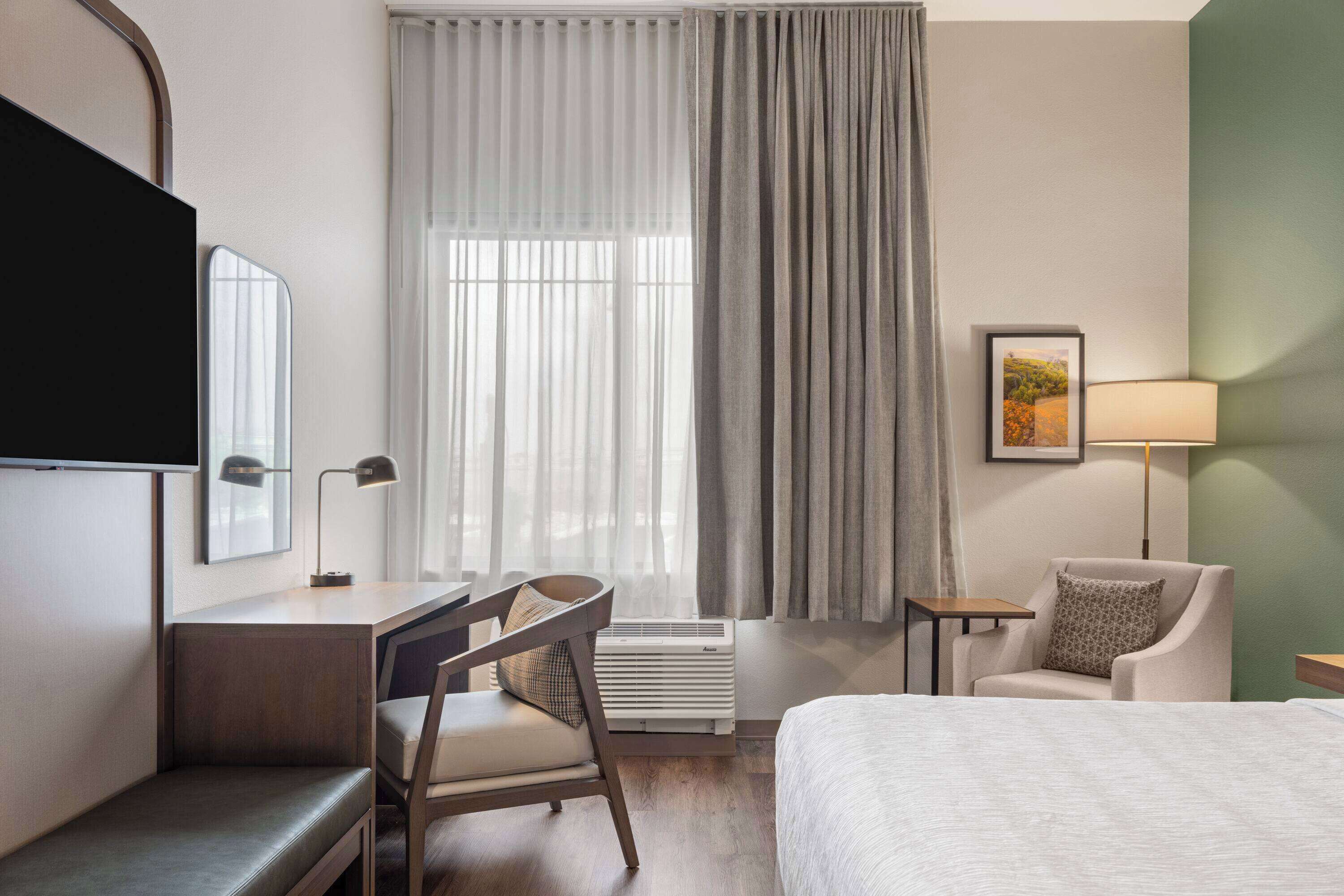 A hotel room with a bed, a desk with a chair, a TV, a lamp, and a window with curtains.