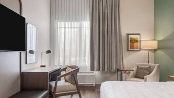 A hotel room with a bed, a desk with a chair, a TV, a lamp, and a window with curtains.