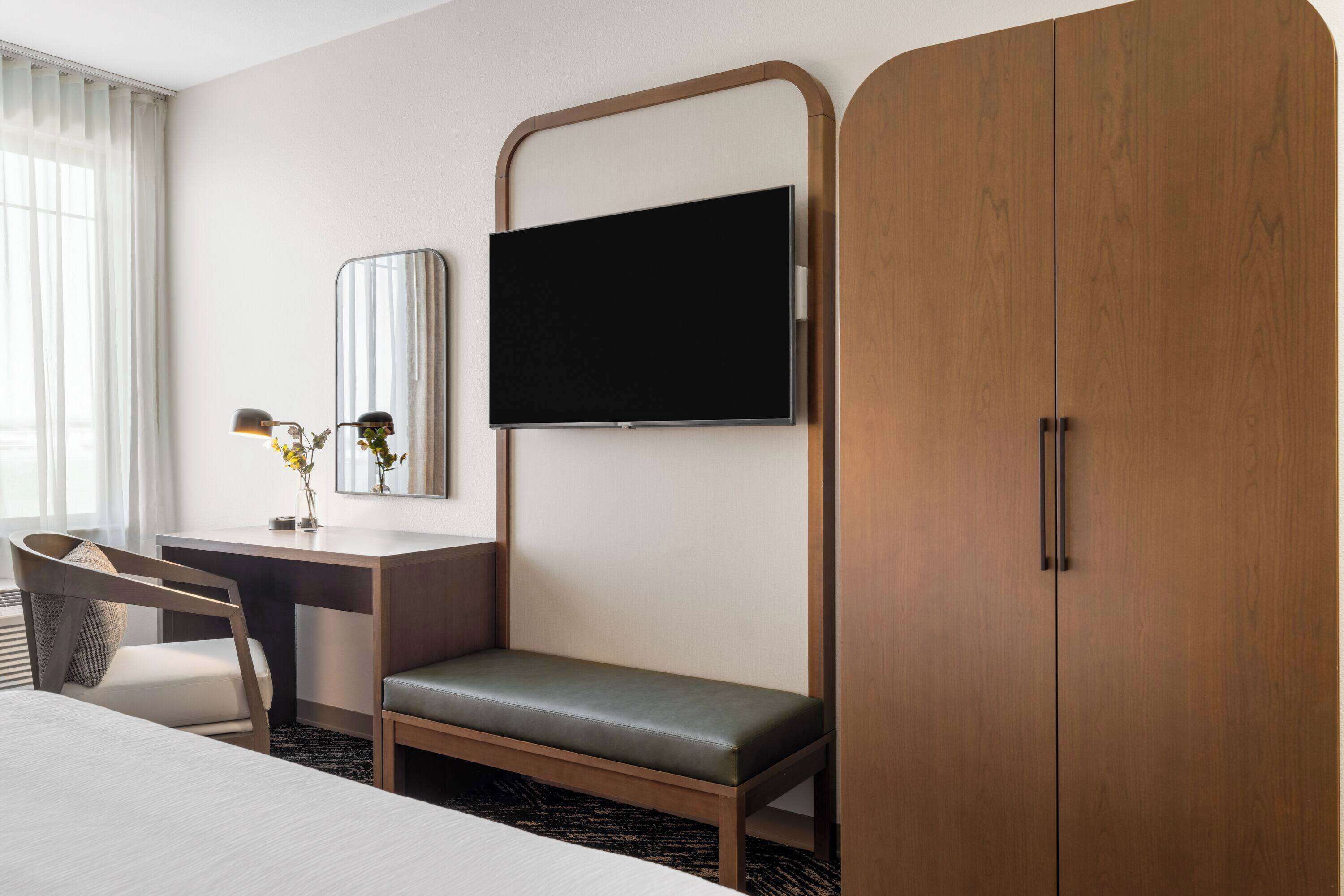 A hotel room with a bed, a desk with a chair, a TV mounted on the wall, and a wardrobe.