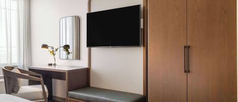 A hotel room with a bed, a desk with a chair, a TV mounted on the wall, and a wardrobe.
