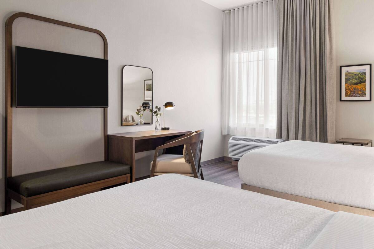 Une chambre d’hôtel comprenant un lit, un bureau avec une chaise, un téléviseur à écran plat, un miroir, une lampe et une fenêtre avec des rideaux.