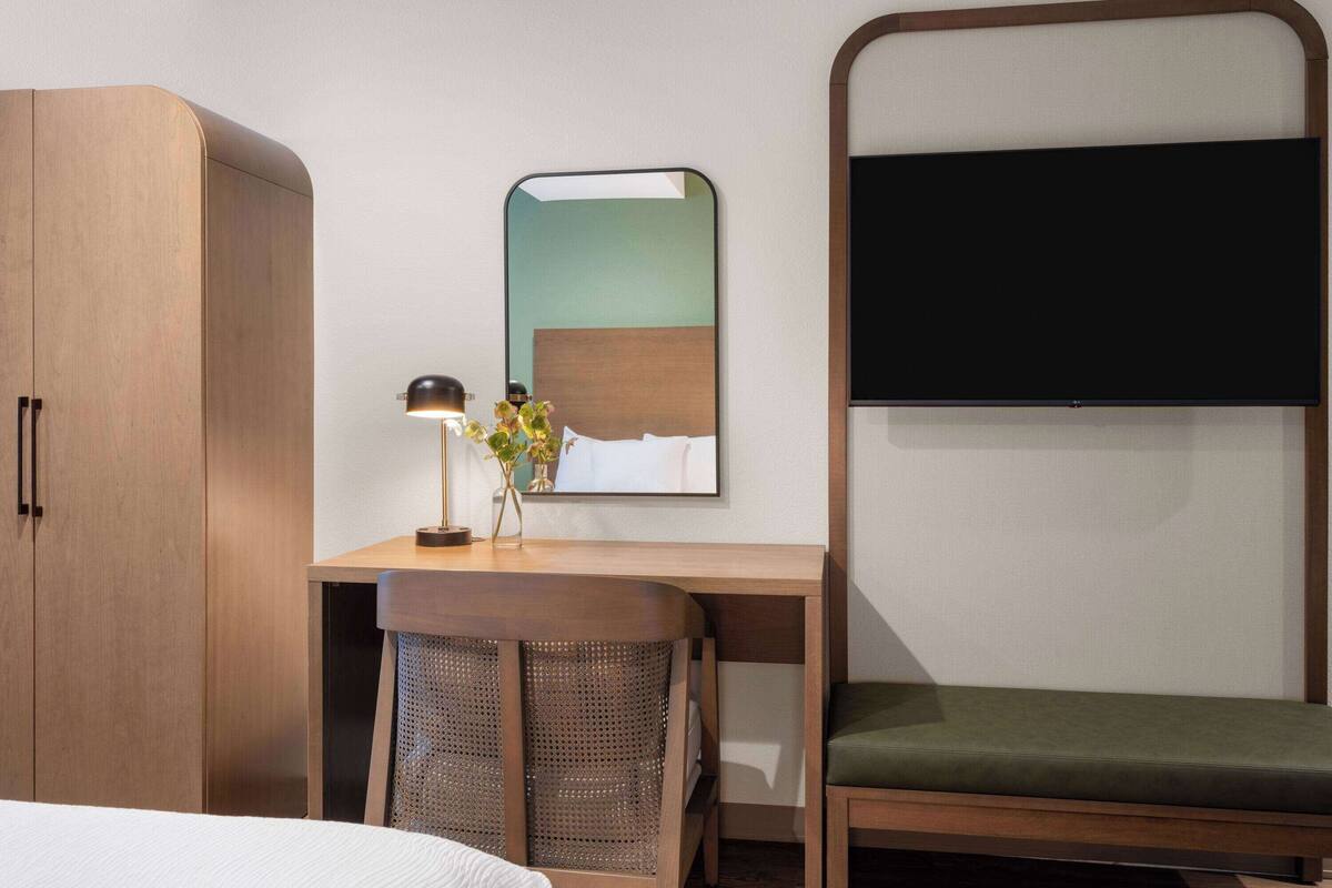 Une chambre d’hôtel avec une armoire en bois, un bureau avec une lampe, un miroir, une télévision et un banc recouvert d’un tissu vert.