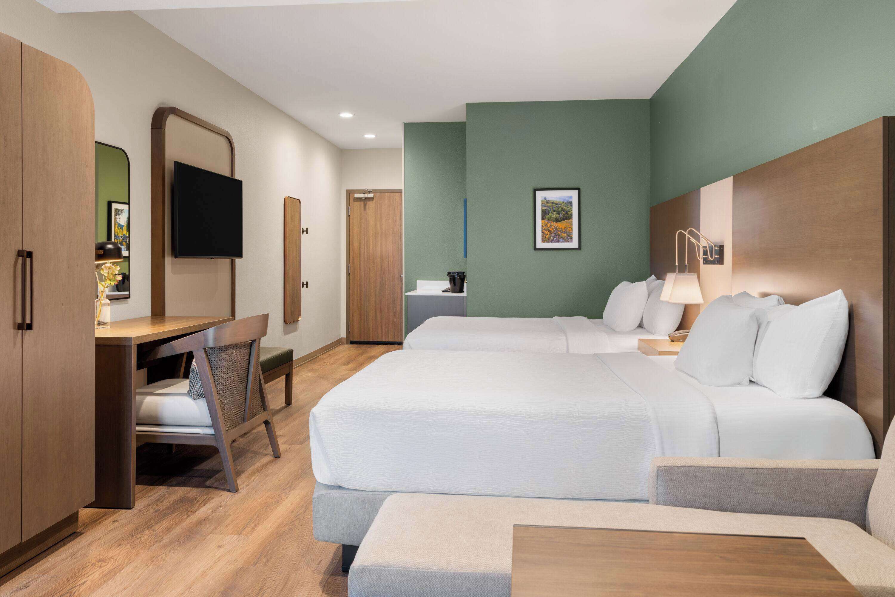 Una camera d'albergo con due letti, una scrivania, una sedia, una TV e un quadro appeso al muro.