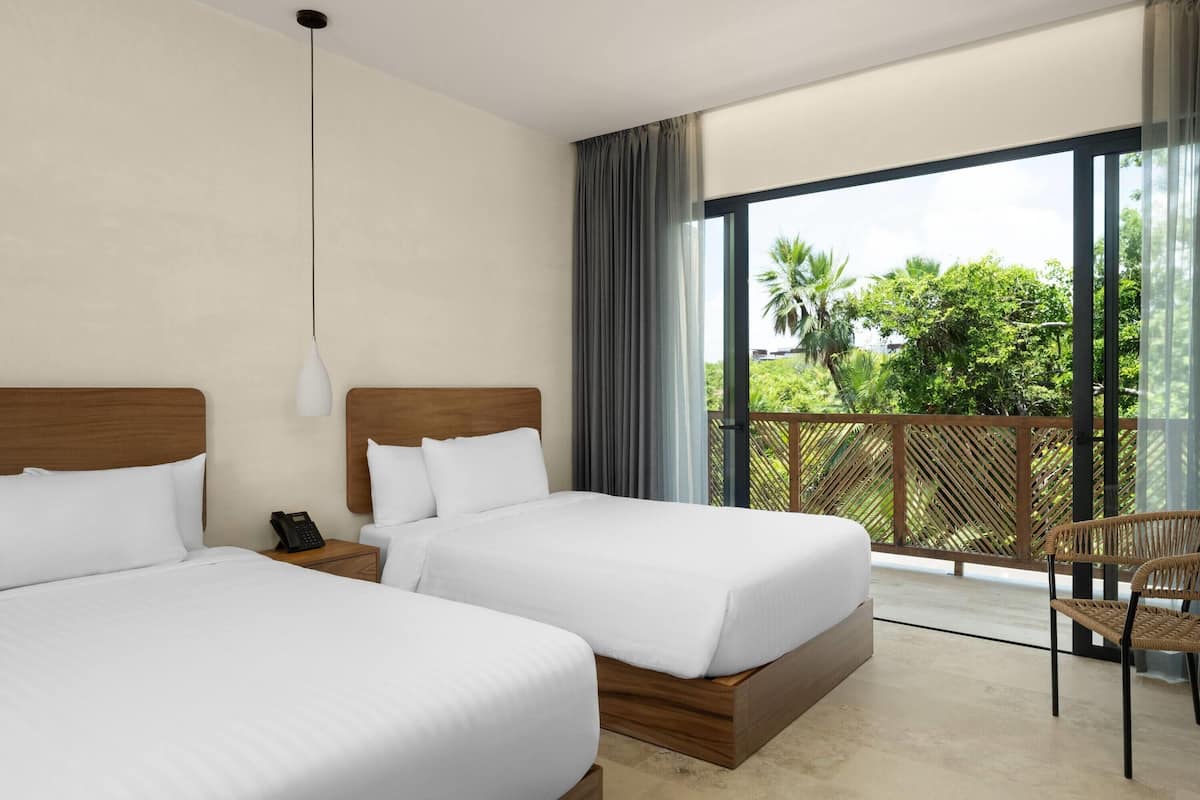 A hotel room with two beds, a wooden headboard, a telephone, and a balcony with a view of greenery.