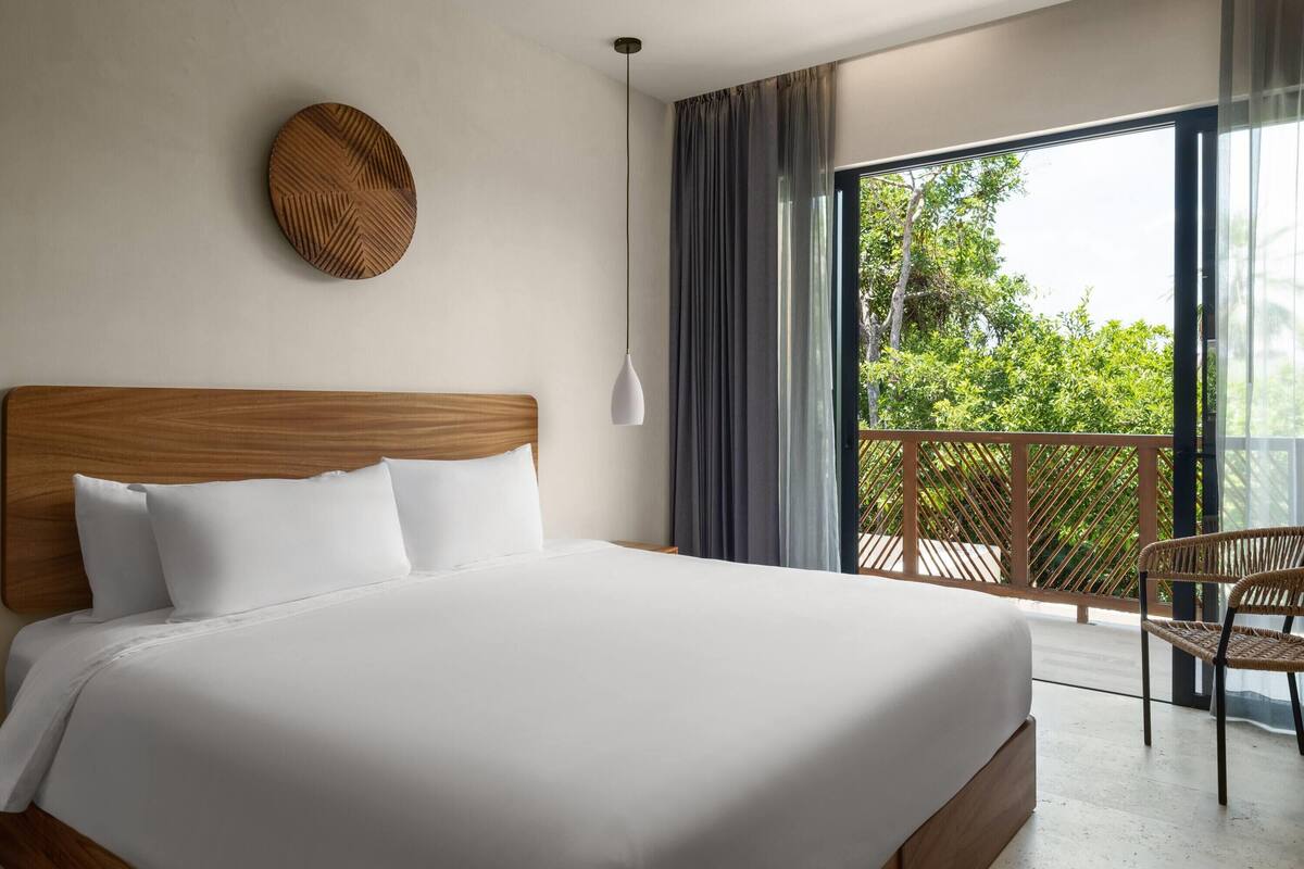 A hotel room with a large bed, a wooden headboard, a round wall decoration, a hanging light, and a view of greenery through a balcony.