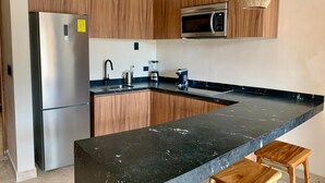 A modern kitchen with a stainless steel refrigerator, a built-in microwave, wooden cabinets, a black granite countertop, and a central island with wooden stools. - Wyndham Residences Tulum (Tulum)