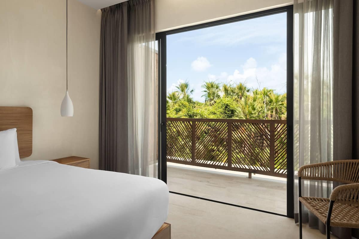 A hotel room with a bed, a nightstand, a wicker chair, and a balcony with a view of palm trees.