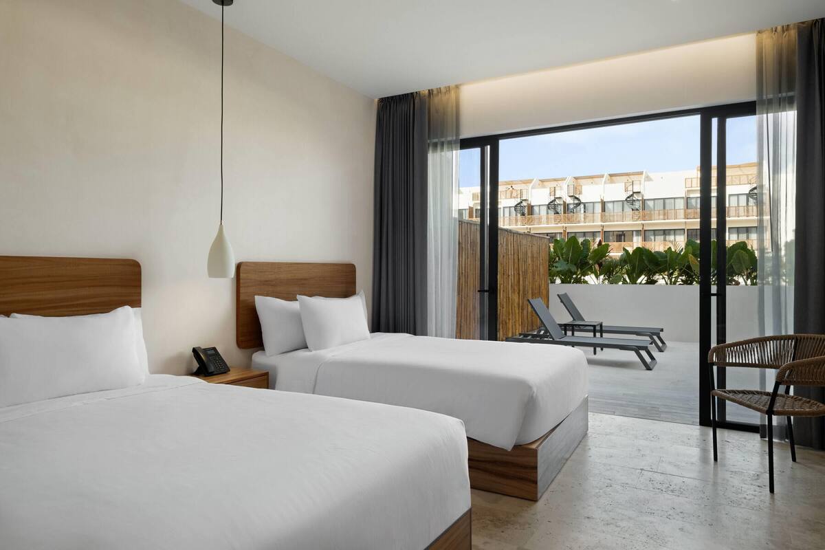 A hotel room with two beds, a phone on the nightstand, a balcony with lounge chairs, and a view of buildings.