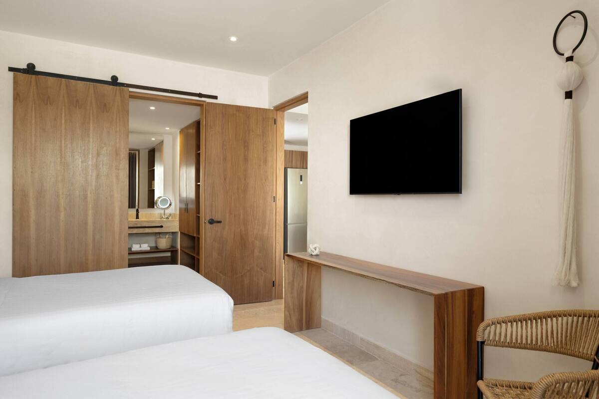 A modern hotel room with two beds, a wooden wardrobe, a vanity with a mirror, a flat-screen TV mounted on the wall, and a wicker chair.
