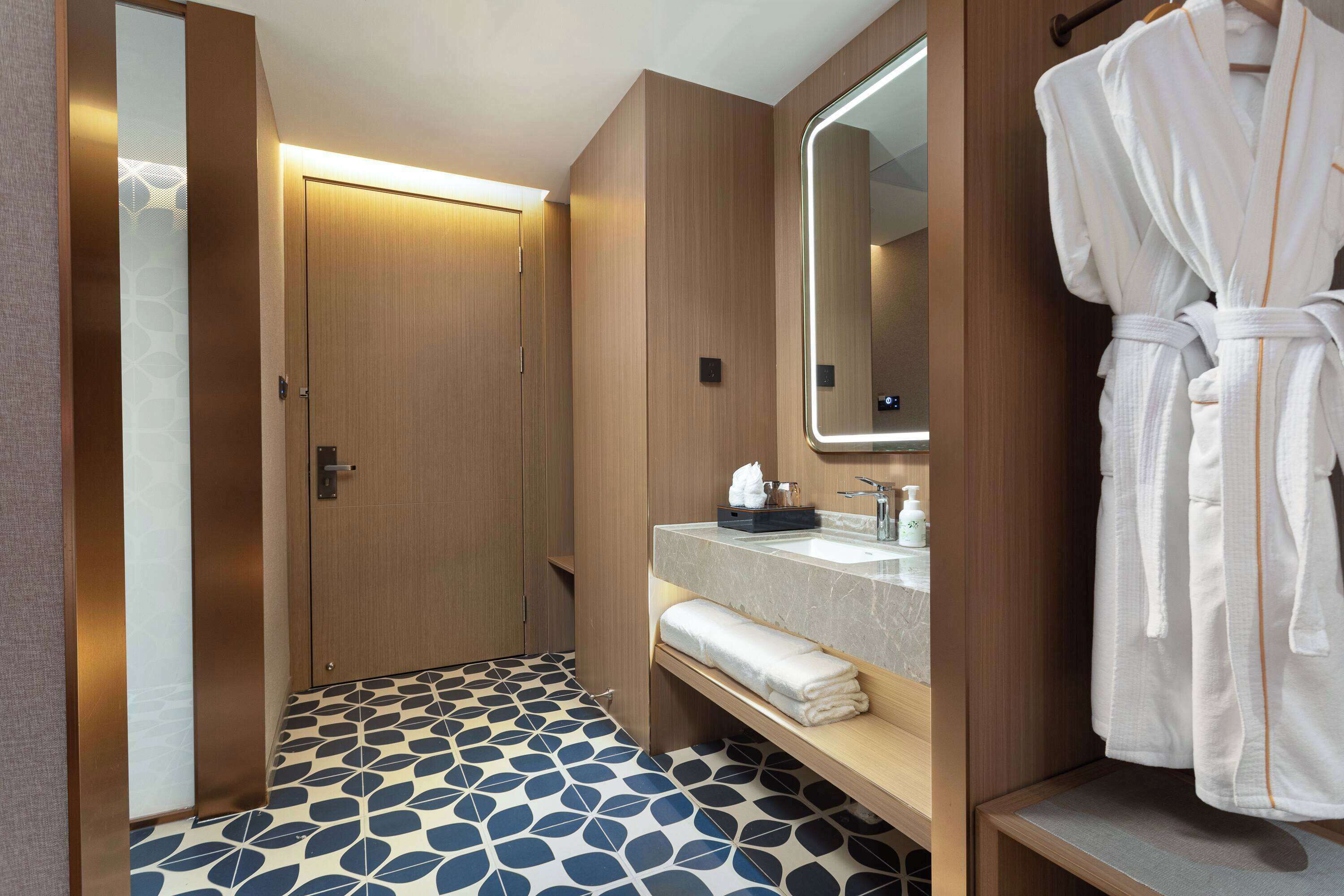 A hotel bathroom with a wooden vanity, a large mirror, a robe hanging on the wall, and a patterned floor.