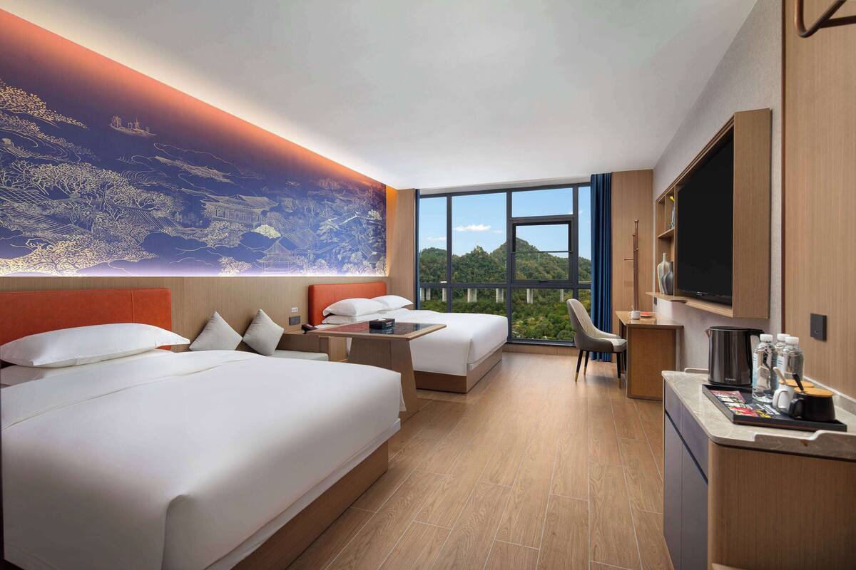 Ein Hotelzimmer mit zwei Betten, einem Schreibtisch, einem Sessel, einem Fernseher und einem großen Fenster mit Blick auf Grünflächen.