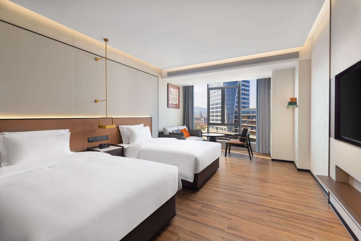 A hotel room with two beds, a desk, a chair, a TV, and a large window with a city view.