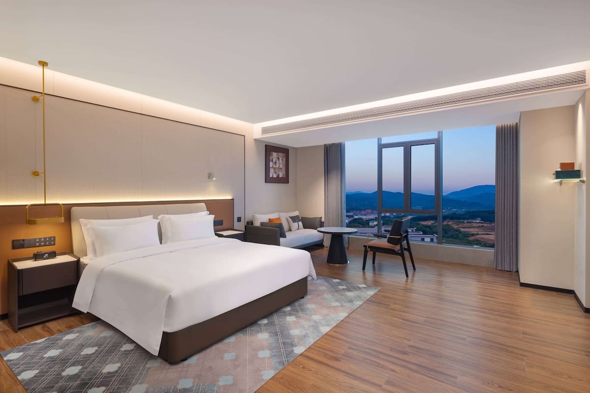A modern hotel room with a large bed, a desk, a sofa, and a view of the cityscape.