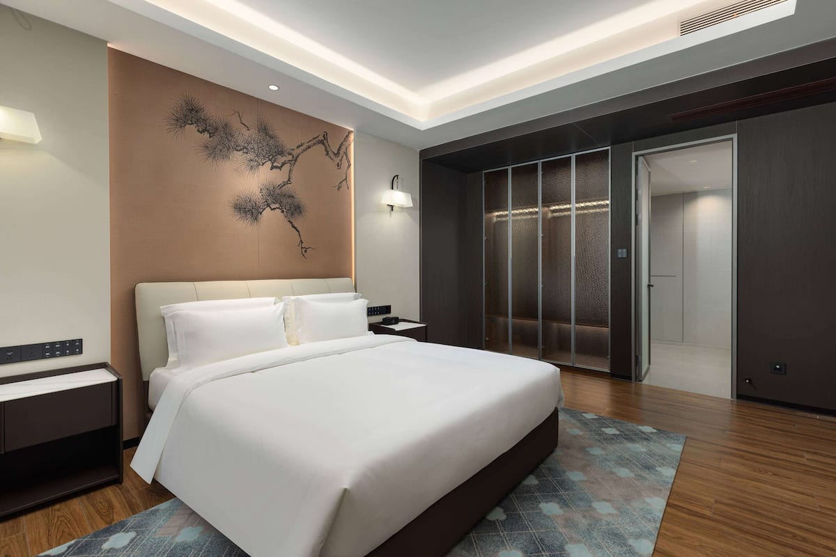A modern hotel room with a large bed, a wall-mounted artwork, a wardrobe, and a bedside table.