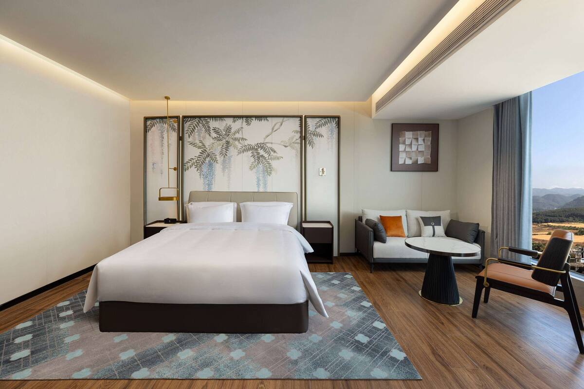 A modern hotel room with a large bed, a sofa, a small table, and a view of the mountains.