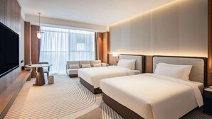 Ein Hotelzimmer mit zwei Betten, einem Fernseher, einem Sofa, einem kleinen Tisch und einem großen Fenster mit Vorhängen.