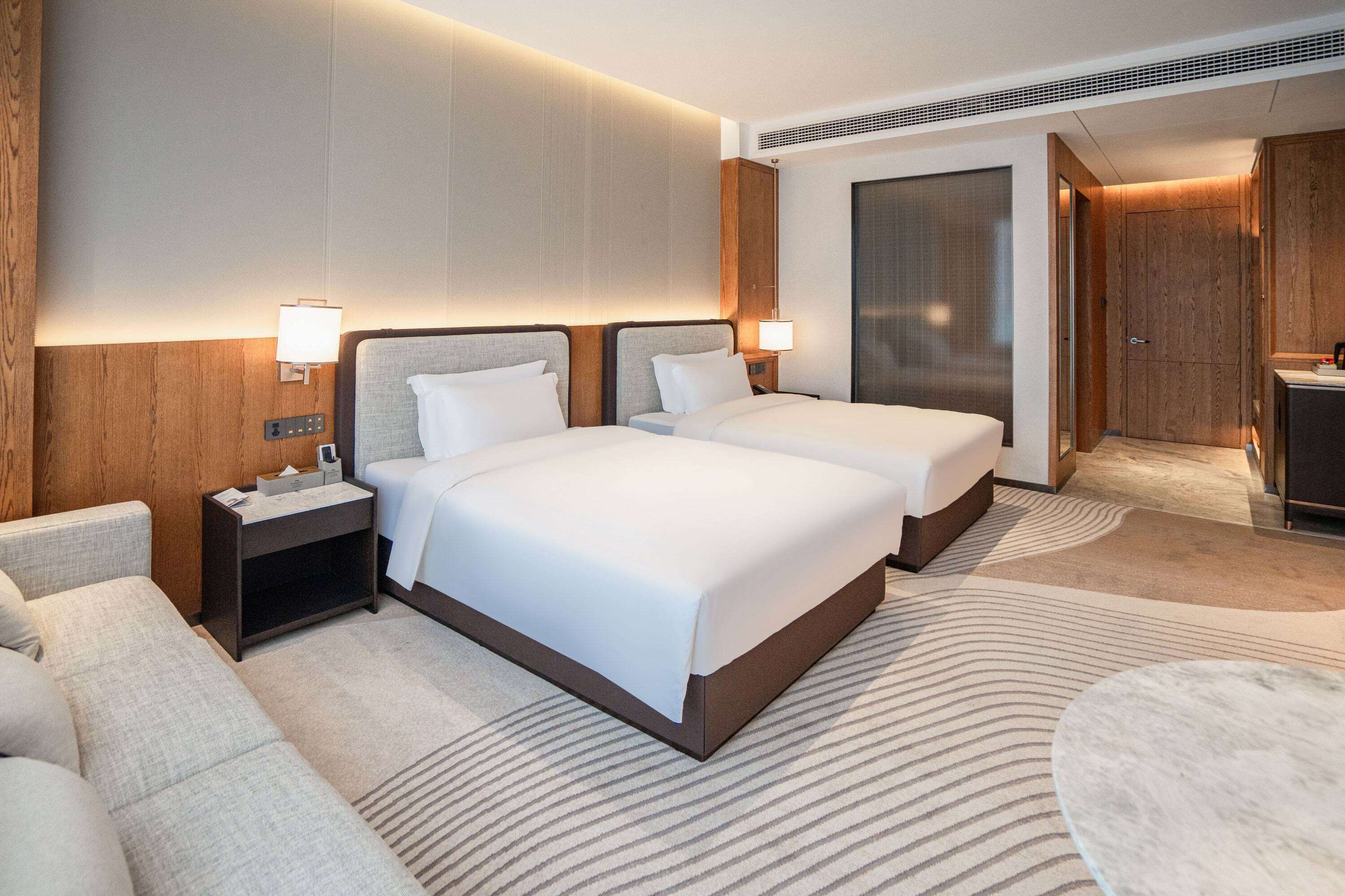 Quarto de hotel com duas camas, um sofá, uma mesa pequena e uma escrivaninha com abajur.
