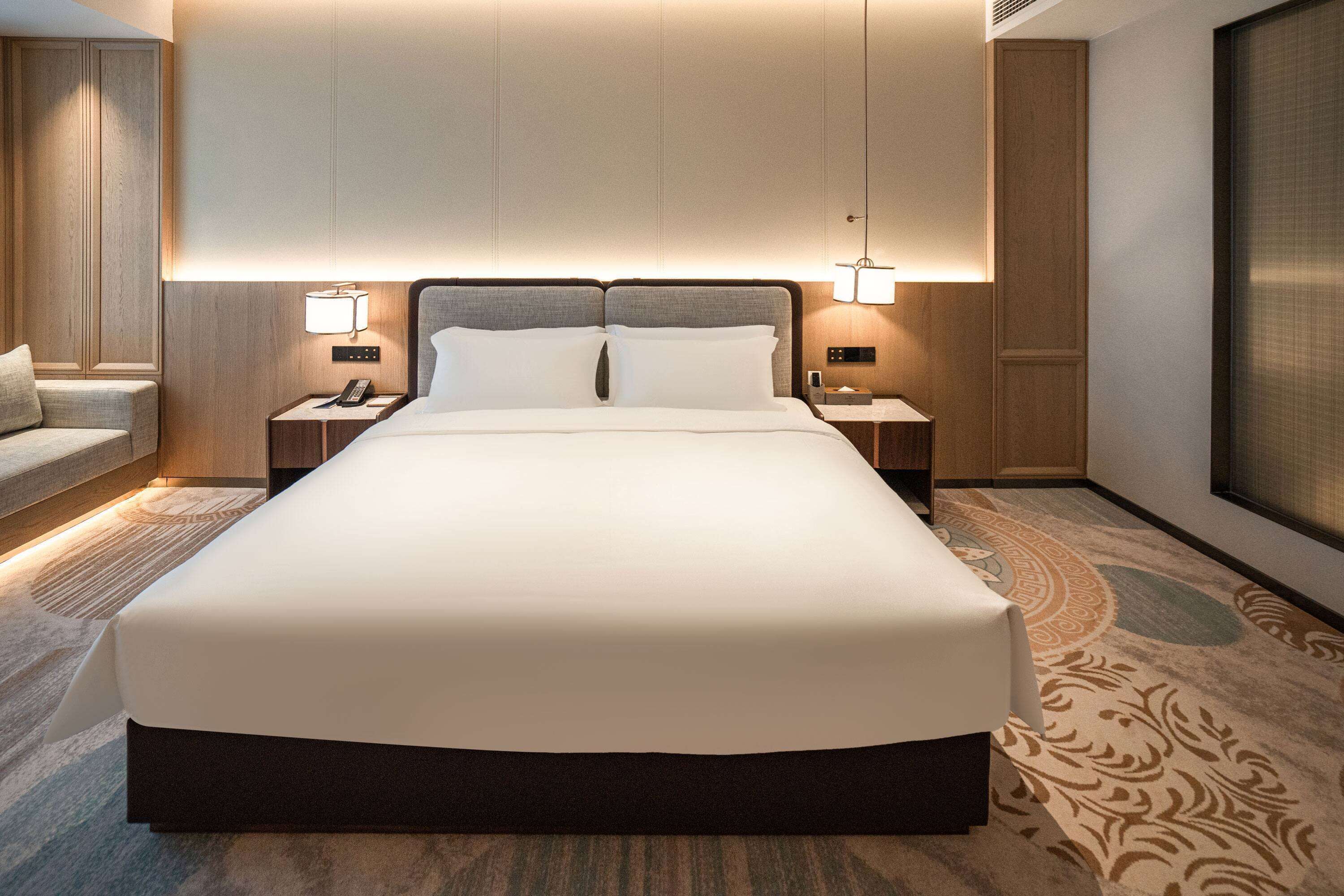 Ein modernes Hotelzimmer mit einem großen Bett, zwei Nachttischen inklusive Lampen, einer kleinen Sitzecke und einem großen Fenster.