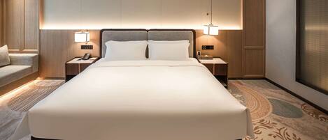 A modern hotel room with a large bed, two bedside tables with lamps, a small seating area, and a large window.