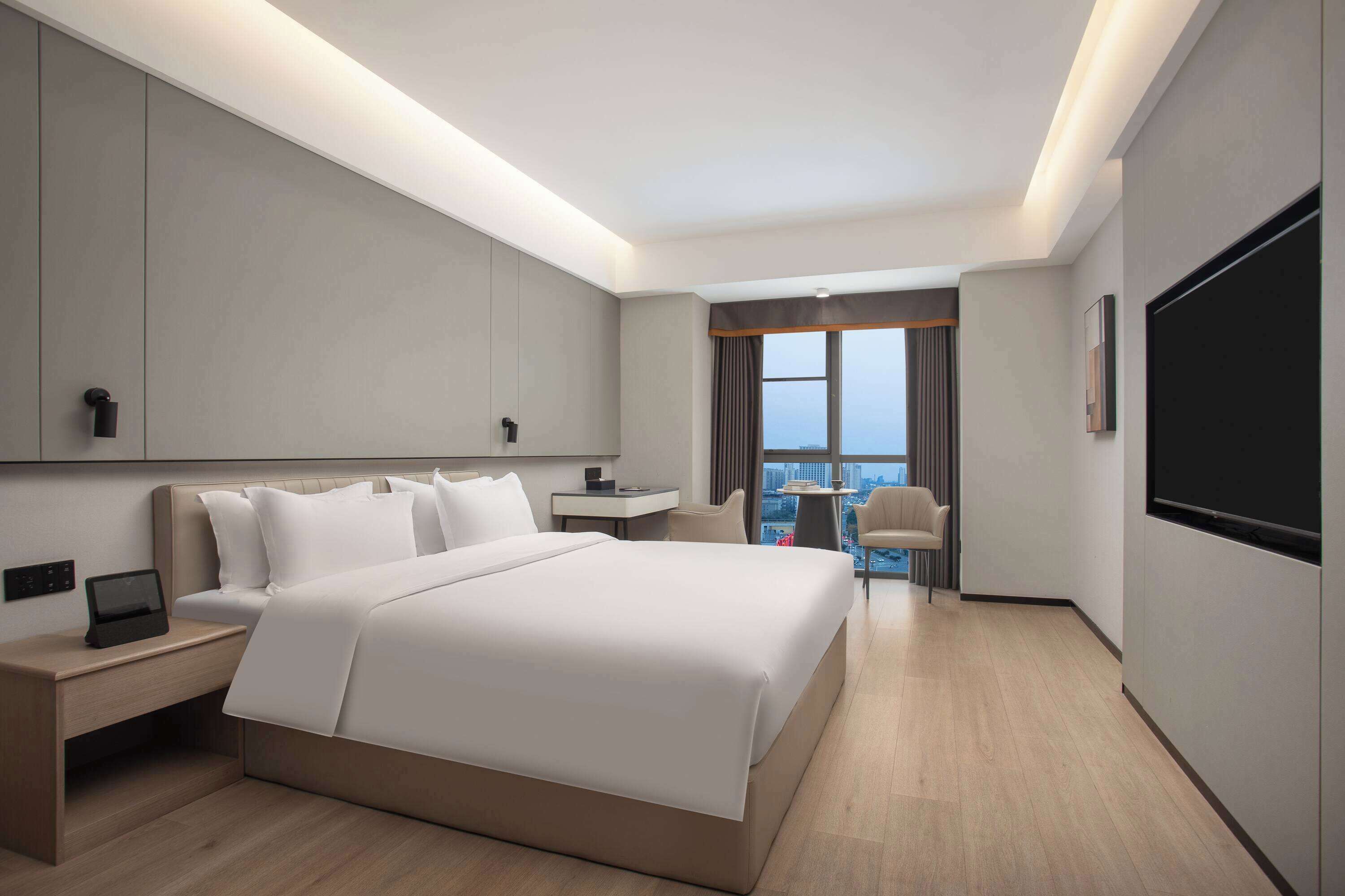 A modern hotel room with a large bed, bedside tables, a desk, and a view of the city through a window.