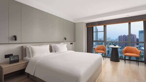 A modern hotel room with a large bed, a bedside table with a radio, two orange chairs around a small table, and a city view through large windows.