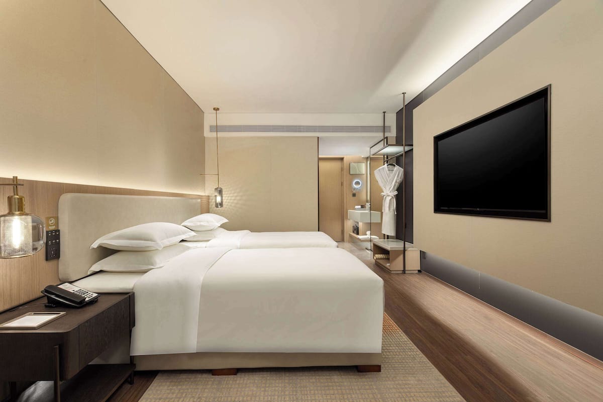 A modern hotel room with two beds, a flat-screen TV, a bedside table with a lamp, and a mirror.