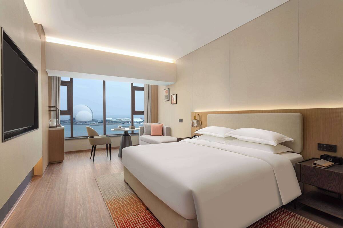 A hotel room with a large bed, a desk, and a view of the cityscape through large windows.