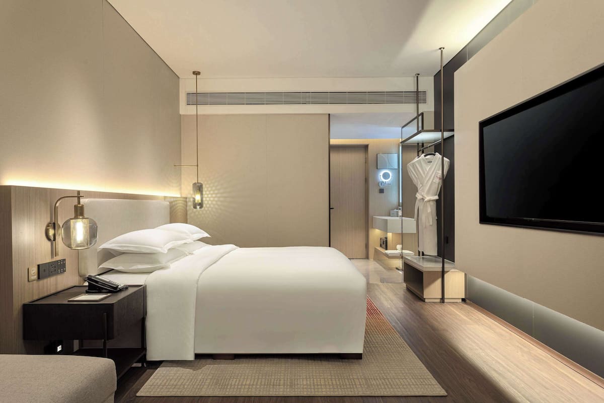 A modern hotel room with a large bed, bedside table, lamp, and a TV mounted on the wall.