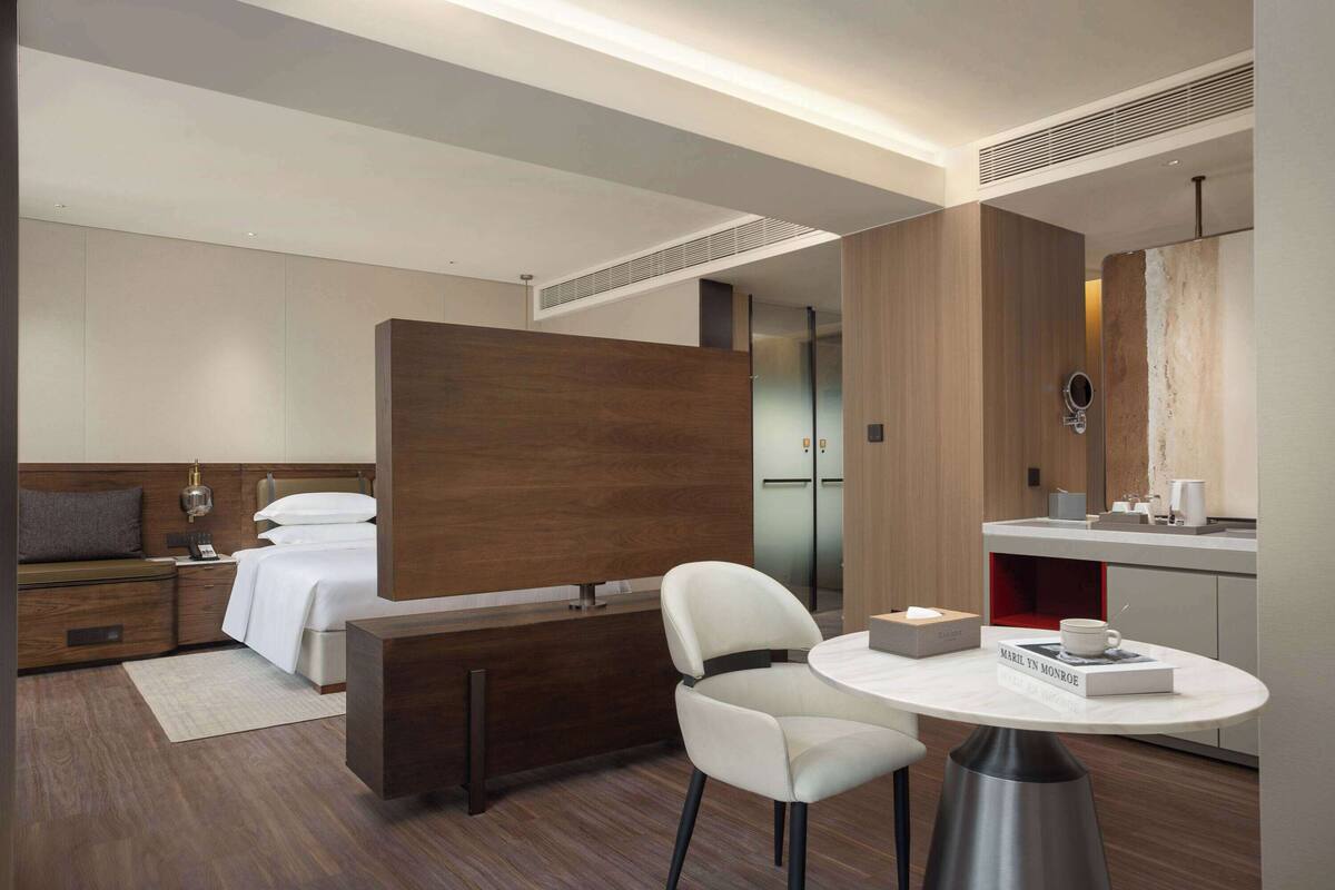 A modern hotel room with a bed, a desk, a chair, a round table, and a mirror.