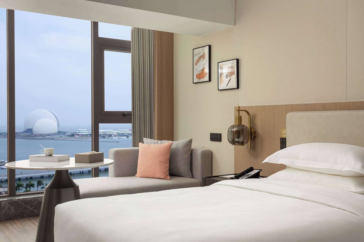 A hotel room with two beds, a small table with a cup and box, a phone on the nightstand, and a view of a cityscape and a large building through the window.