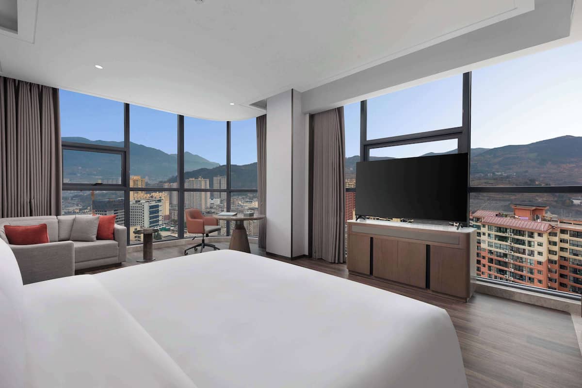 A modern hotel room with a large bed, a TV, a desk, and a view of the city and mountains.