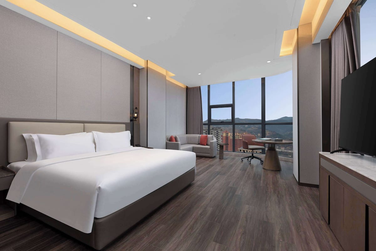 A modern hotel room with a large bed, a desk, a chair, and a TV. The room has a city view.