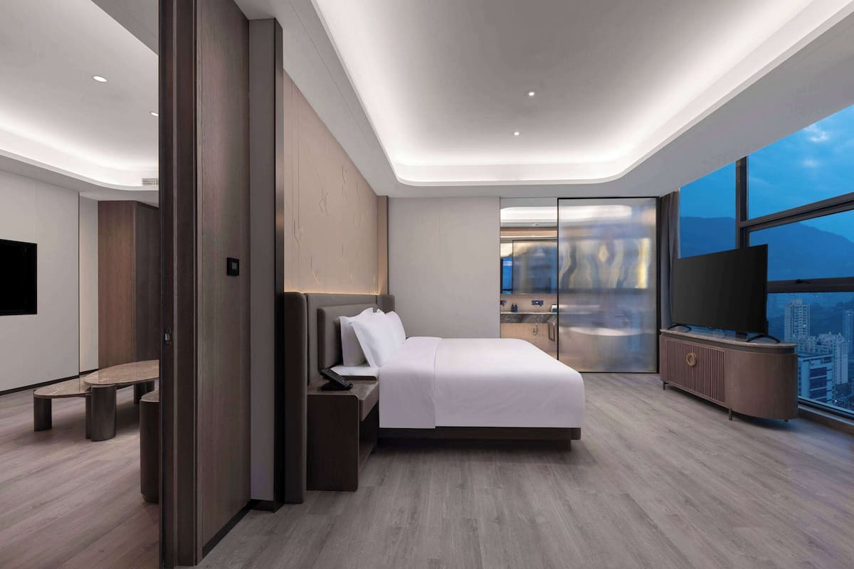 A modern hotel room with a bed, bedside table, and a city view through large windows.
