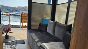 Ein modernes Interieur mit einem grauen Sofa, einem kleinen runden Tisch mit einer Vase und Blick auf einen Steg mit Booten.